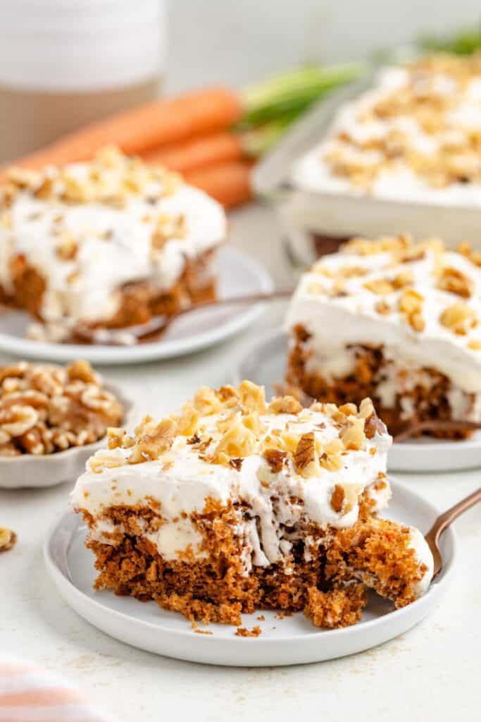 Sliced carrot poke cake with cream cheese frosting, topped with nuts; fork and fresh carrots nearby.