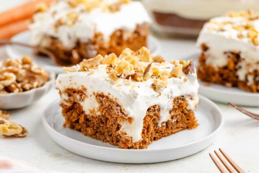 Slice of carrot poke cake with frosting and walnuts on plate; additional slices and walnuts in background.