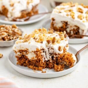 Slice of frosted carrot poke cake with walnuts on a plate; fork taking a bite.