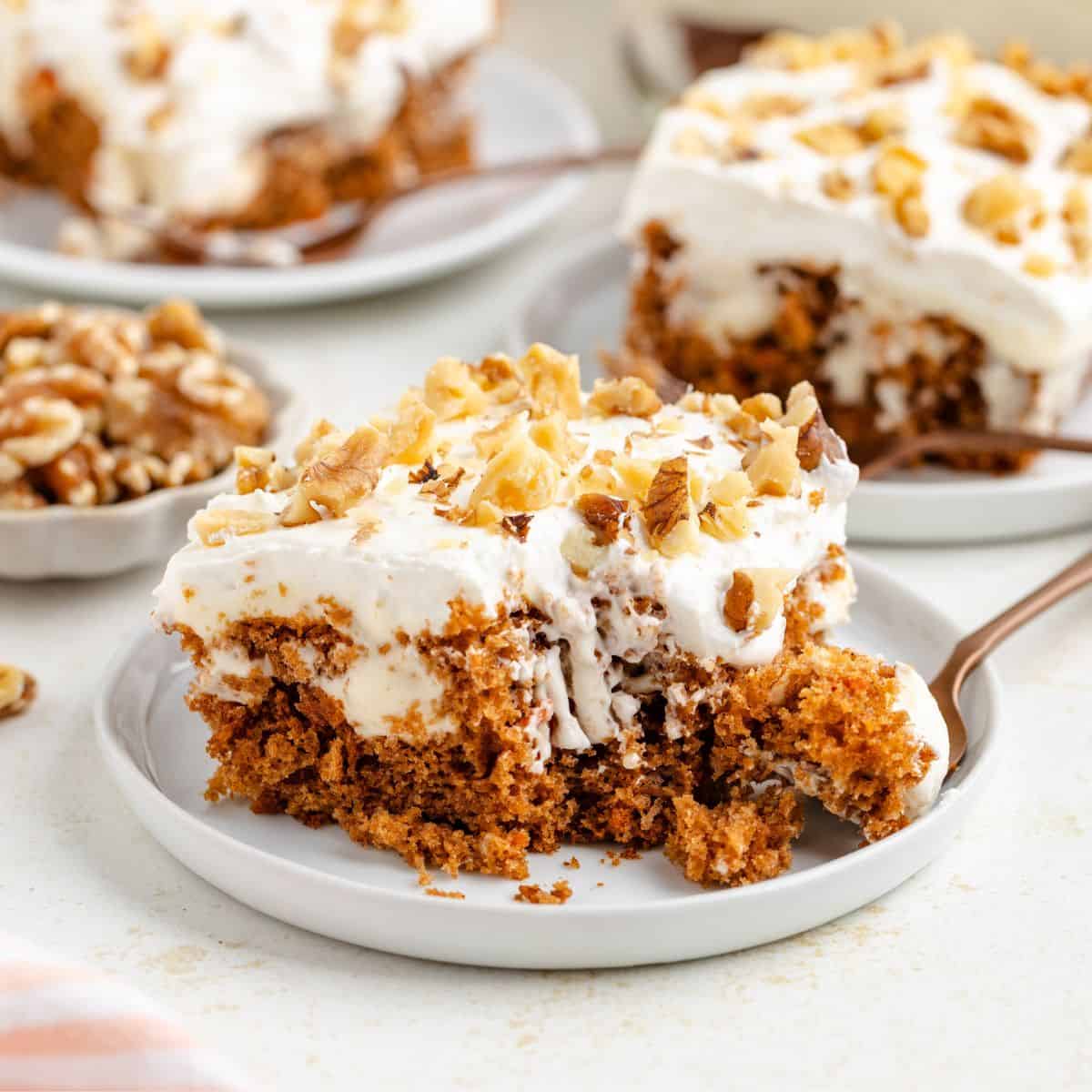 Slice of frosted carrot poke cake with walnuts on a plate; fork taking a bite.