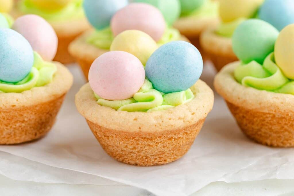 Easter cookie cups topped with green frosting and pastel-colored candy eggs on a sheet of parchment paper.