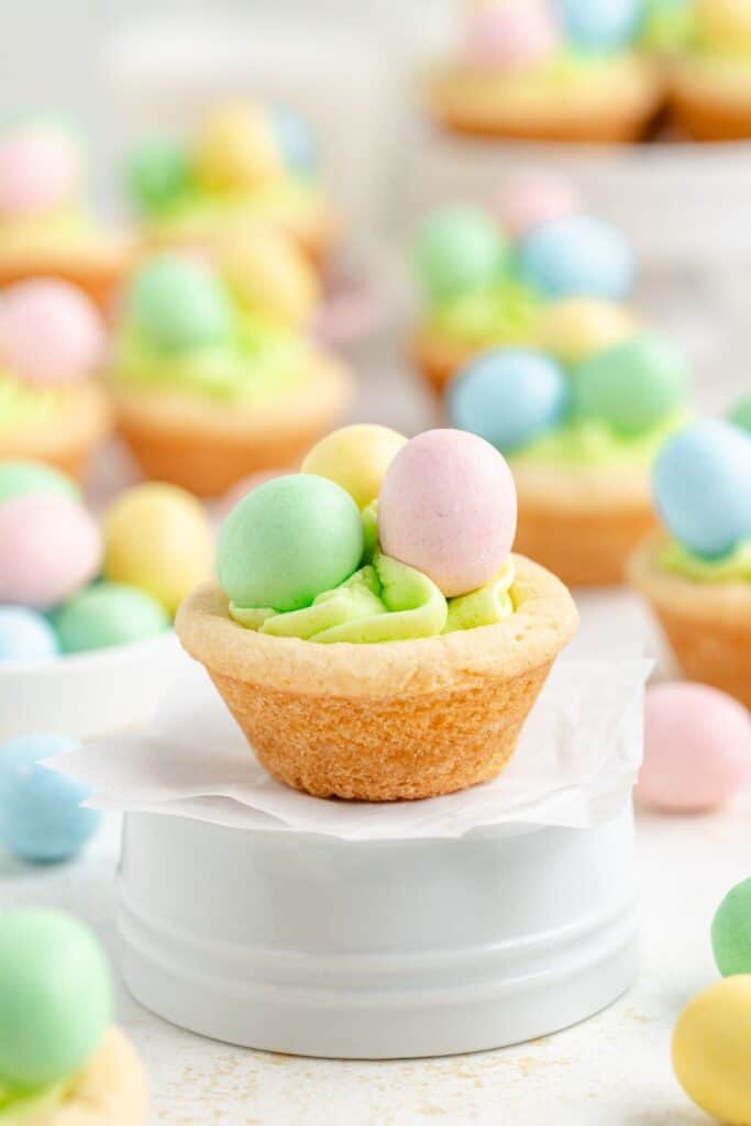 A cookie cup filled with green frosting and topped with pastel-colored chocolate eggs, placed on white parchment paper over a white surface. More decorated cookie cups are blurred in the background.