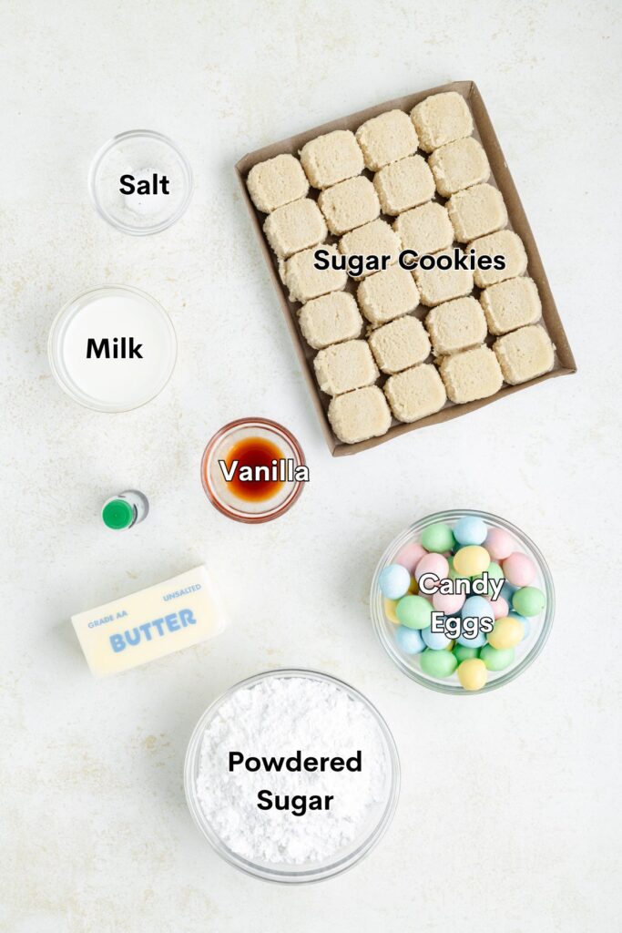 Overhead view of ingredients for decorating sugar cookies: sugar cookies, milk, salt, vanilla, candy eggs, powdered sugar, butter, and green food coloring.