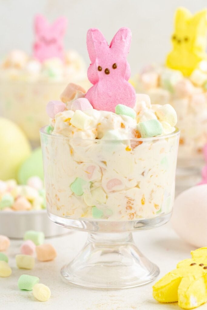 Dessert cup with colorful marshmallows and cream, topped by pink chick, evokes Easter charm; yellow chick behind.