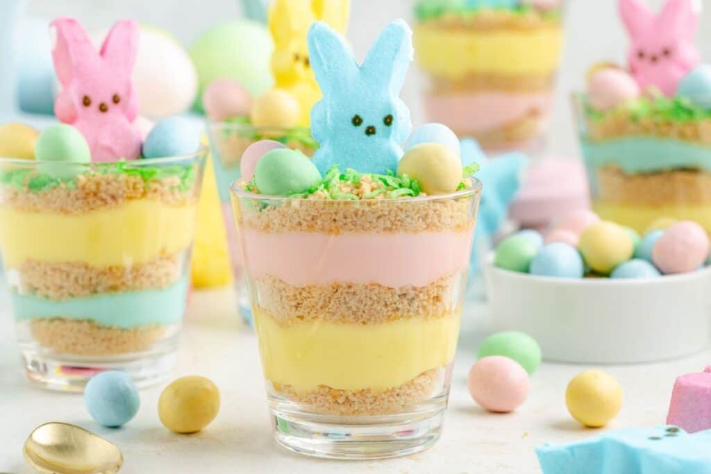 Easter pudding cup with a blue marshmallow bunny and green coconut grass, surrounded by colorful candy eggs.