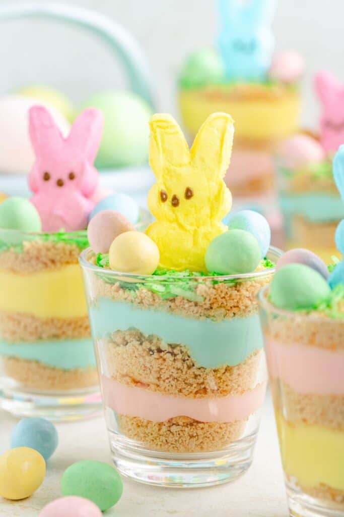 Easter pudding dessert cups with crushed cookies, topped with marshmallow bunnies and candy eggs on a light background.
