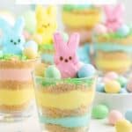 A glass cup with blue, yellow, beige layers, topped by a pink bunny marshmallow and pastel candy eggs.