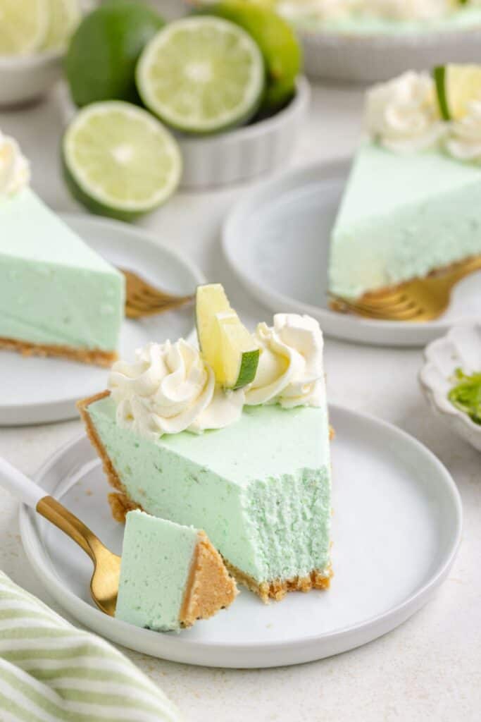 A slice of lime jello pie topped with whipped cream and a lime wedge sits on a white plate with a gold fork. 