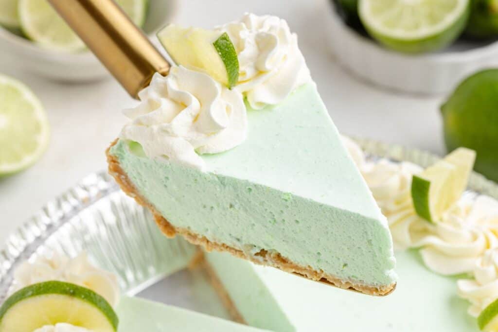 Slice of no-bake lime pie with whipped cream on a golden spatula, topped with lime slices, above a pie tin.