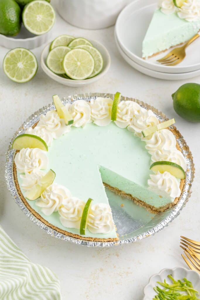 A lime Jello pie topped with whipped cream and lime slices, with one slice missing. Plates and lime halves are in the background.