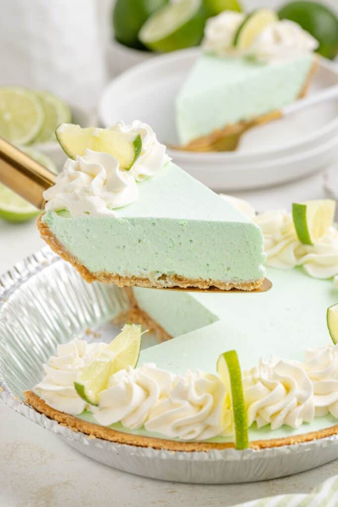 A slice of no-bake lime Jello pie with whipped cream and lime garnish is being lifted from a silver pie dish. 