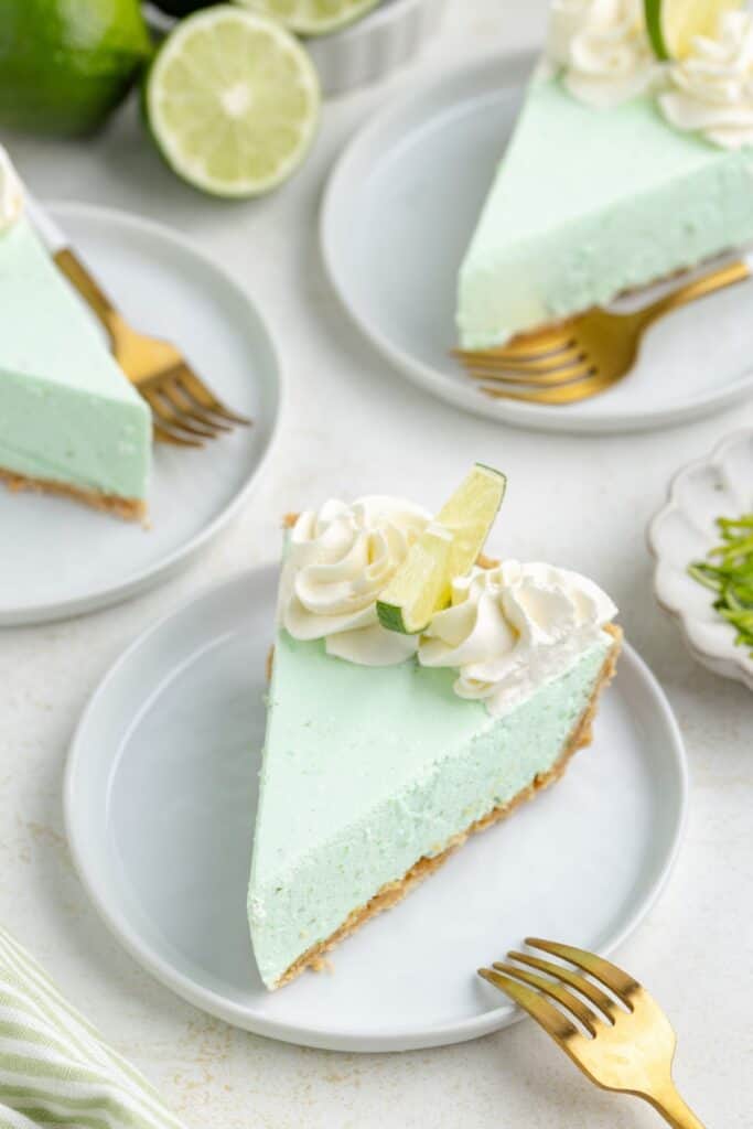 Slices of green key lime Jello pie with whipped cream and lime wedges on white plates, accompanied by gold forks.