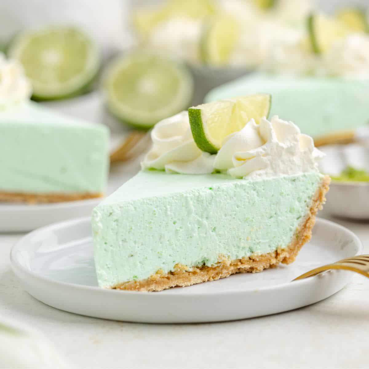 A slice of lime Jello pie with a graham cracker crust, topped with whipped cream and lime wedges, on a white plate.