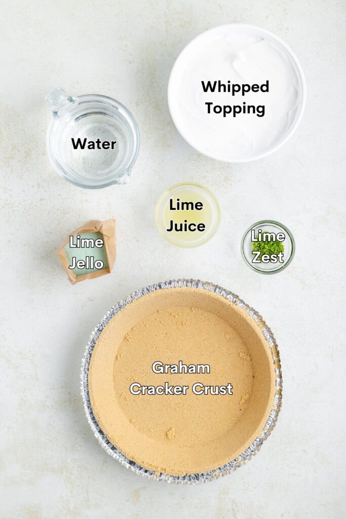 Ingredients for a lime pie include graham cracker crust, whipped topping, lime juice, lime zest, lime Jello, and water, arranged on a light surface.