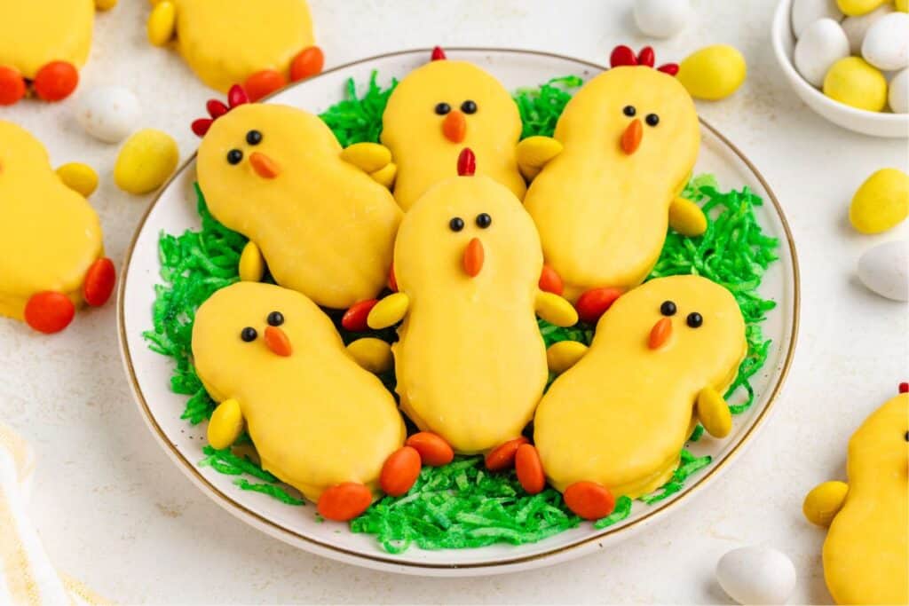 Nutter Butter Easter chick cookies on green coconut grass, surrounded by colorful chocolate eggs.