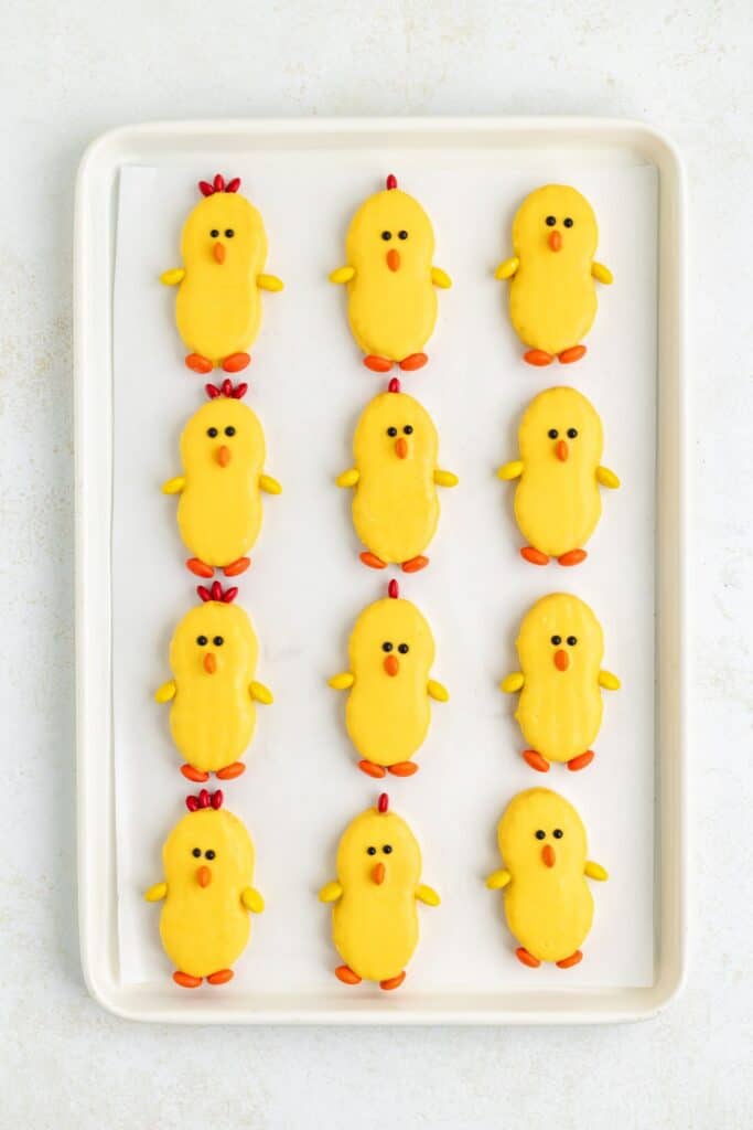 Chick-shaped Nutter Butter cookies with red and orange candy details on a parchment paper-lined baking sheet.