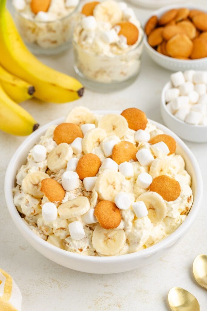 Bowl of banana pudding fluff with sliced bananas, wafer cookies, and marshmallows; whole bananas and wafers surround.