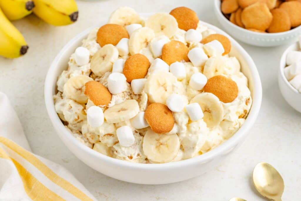 Bowl of banana pudding fluff salad topped with bananas, wafers, and marshmallows; surrounded by bananas.