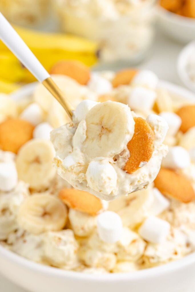 Spoonful of banana pudding with wafers and mini marshmallows held above a matching dessert bowl.