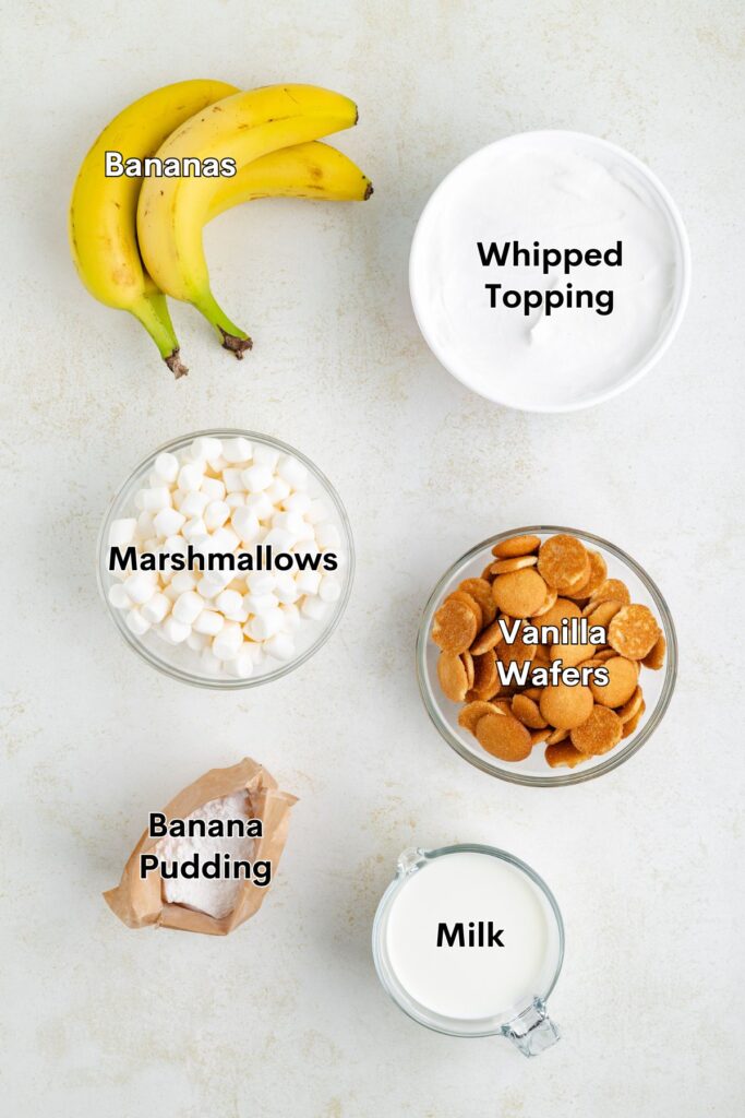 Ingredients for banana pudding: bananas, whipped topping, marshmallows, vanilla wafers, pudding mix, milk on light surface.