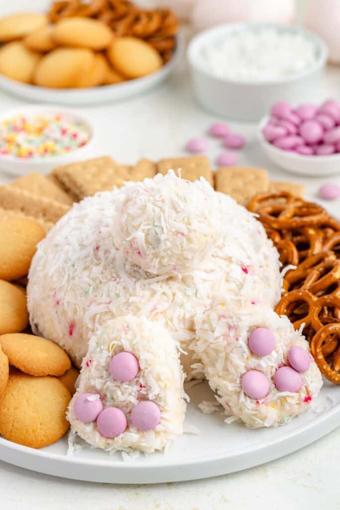 Bunny butt cheese ball covered in coconut flakes, adorned with pink candies; surrounded by pretzels, cookies, crackers.