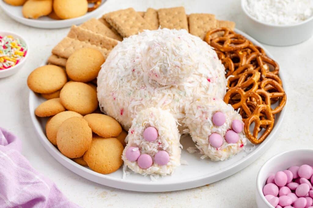 Bunny-shaped cheese ball covered in coconut, with pretzels, crackers, wafers; pink candies for decoration.