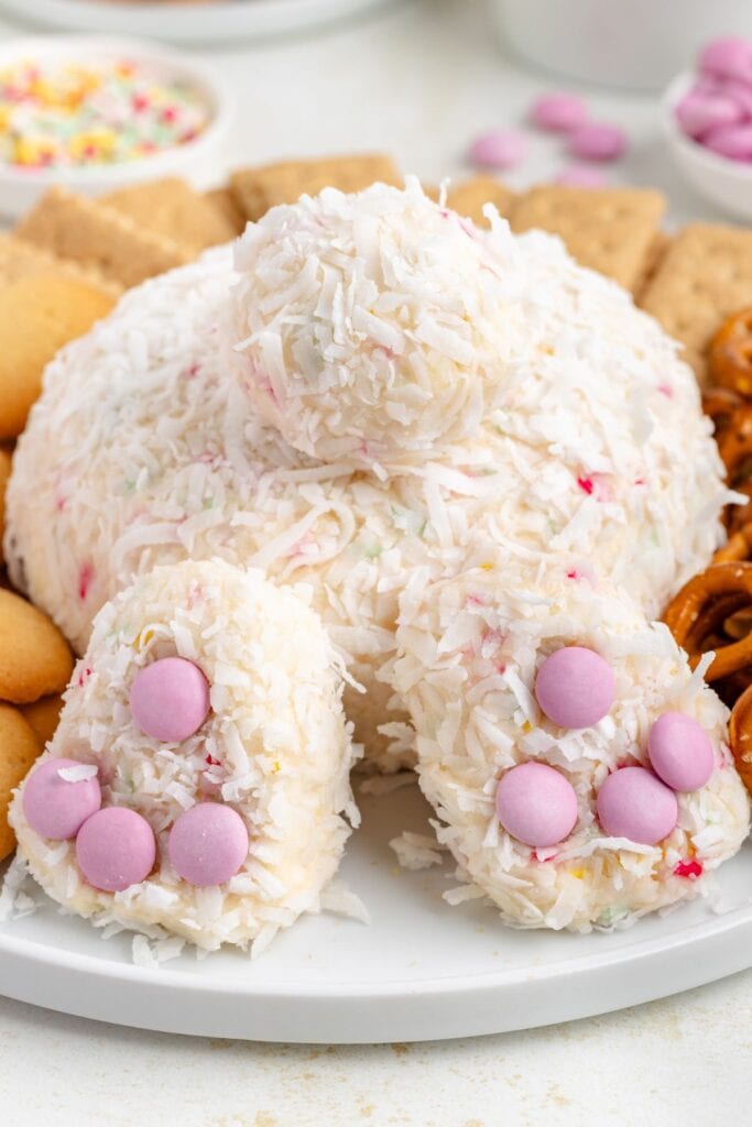 A bunny butt cheese ball dessert covered in coconut, with pink candy feet, surrounded by cookies and pretzels.