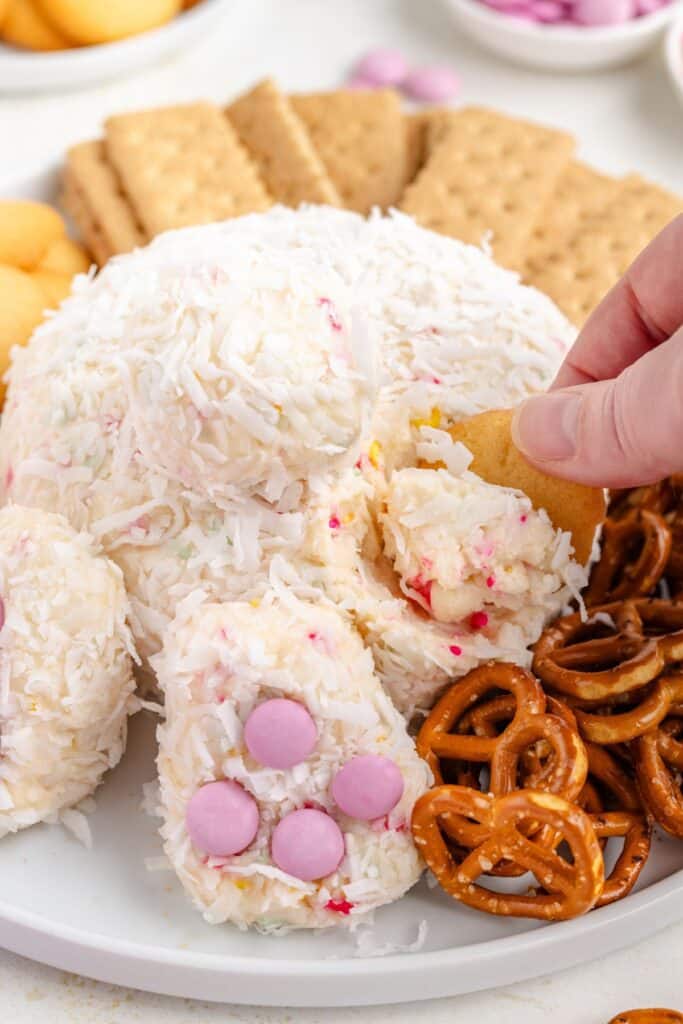 Hand dipping cracker into cheese ball with shredded coconut, pink candies; surrounded by pretzels and graham crackers.