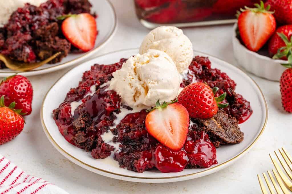 Chocolate strawberry cobbler with vanilla ice cream and fresh strawberries on a dessert plate; extra servings in background.