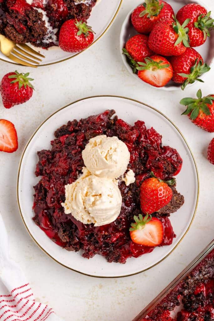 Strawberry chocolate dump cake with two vanilla ice cream scoops, garnished with fresh strawberries and extra servings.