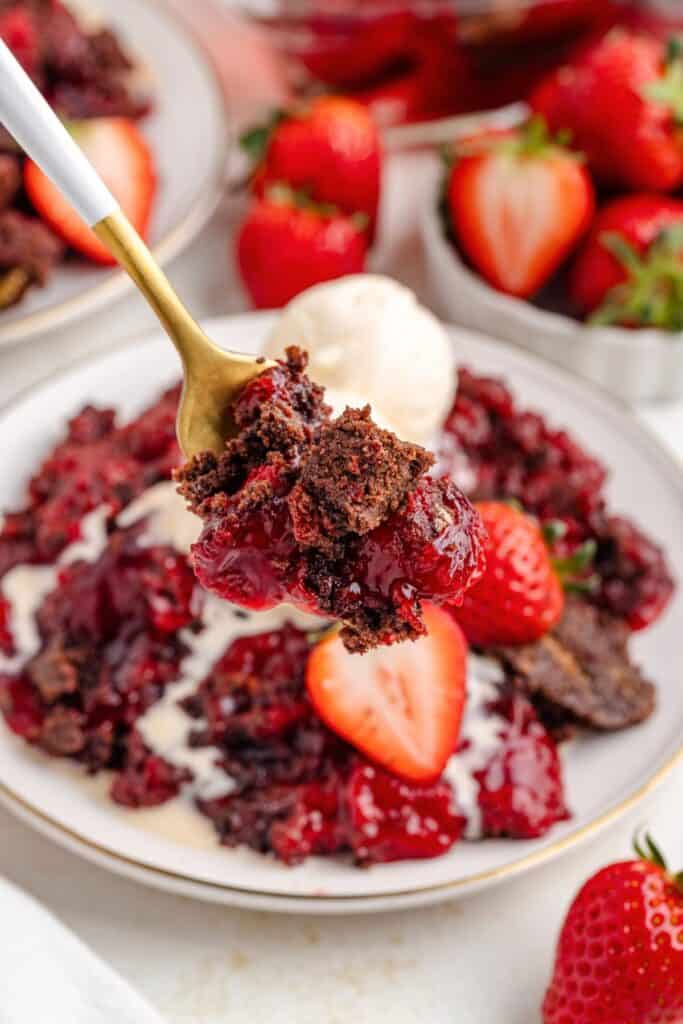 Fork holding strawberry chocolate dessert; plate with more dessert, halved strawberry, and ice cream scoop behind.