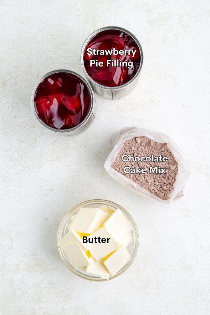 Top-down view of strawberry pie filling cans, chocolate cake mix bag, and bowl of cubed butter on white.