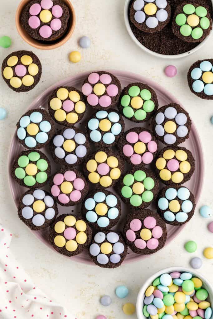 Mini brownie bites with pastel candy flowers on a plate, surrounded by bowls of candies and more cupcakes.