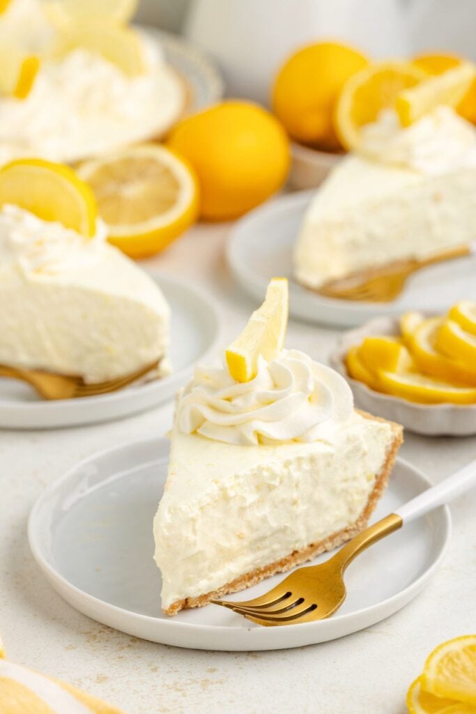 Slice of creamy lemon pie with whipped topping and lemon wedge, on a plate, surrounded by more slices.