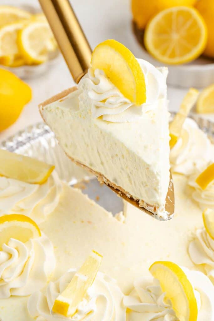Close-up of creamy lemon no-bake pie slice with whipped topping and lemon wedge; whole lemons in background.