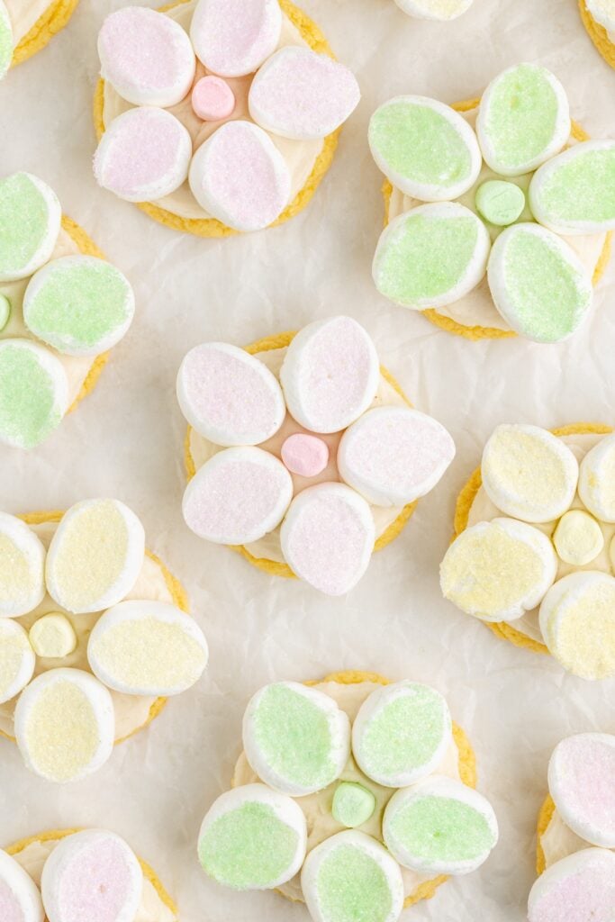 Pastel marshmallow flowers atop frosted cookies on a light background.