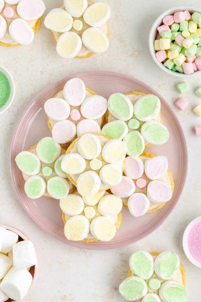 Cookies with pastel marshmallow flowers on pink plate, surrounded by mini marshmallows and colored sugar bowls.