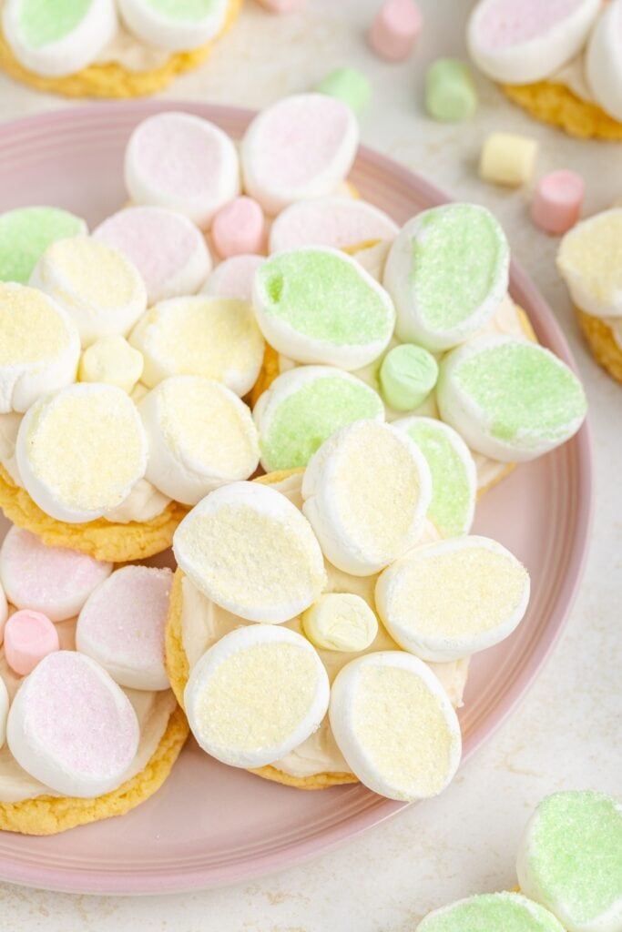 Plate of cookies topped with pastel marshmallow flowers, surrounded by scattered colorful marshmallows.