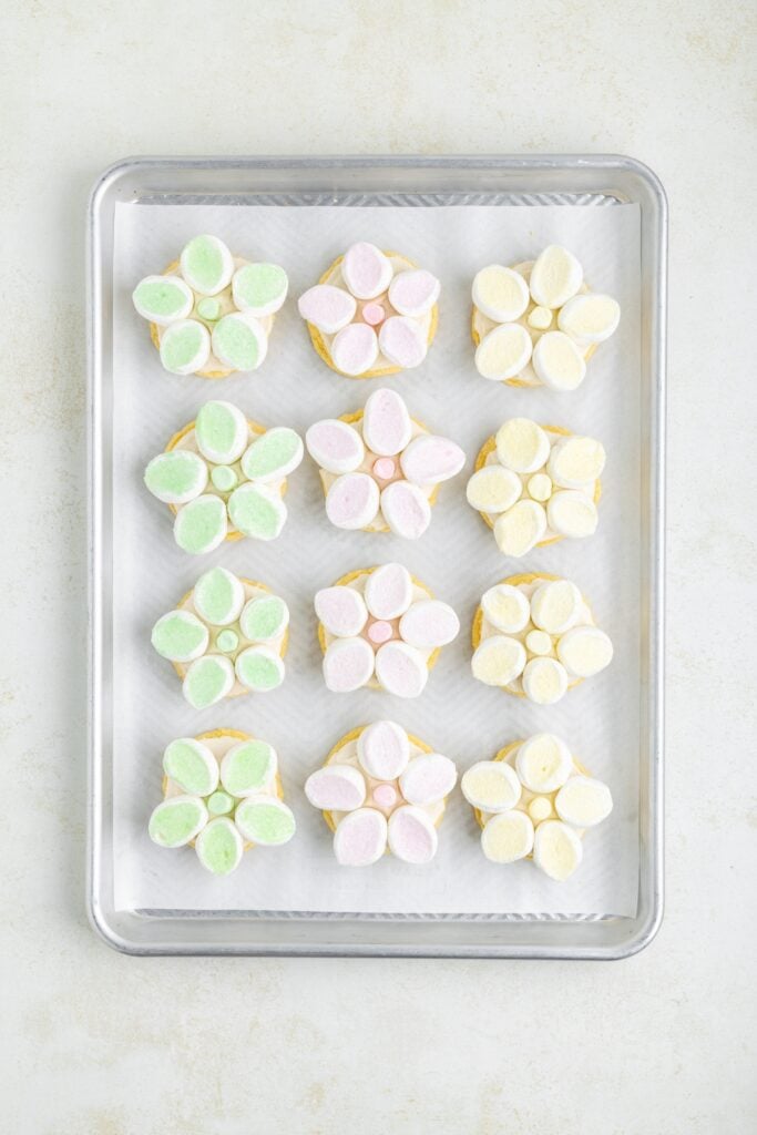 Flower-shaped cookie sandwiches with pastel marshmallows in mint green, yellow, and pink on a baking tray.