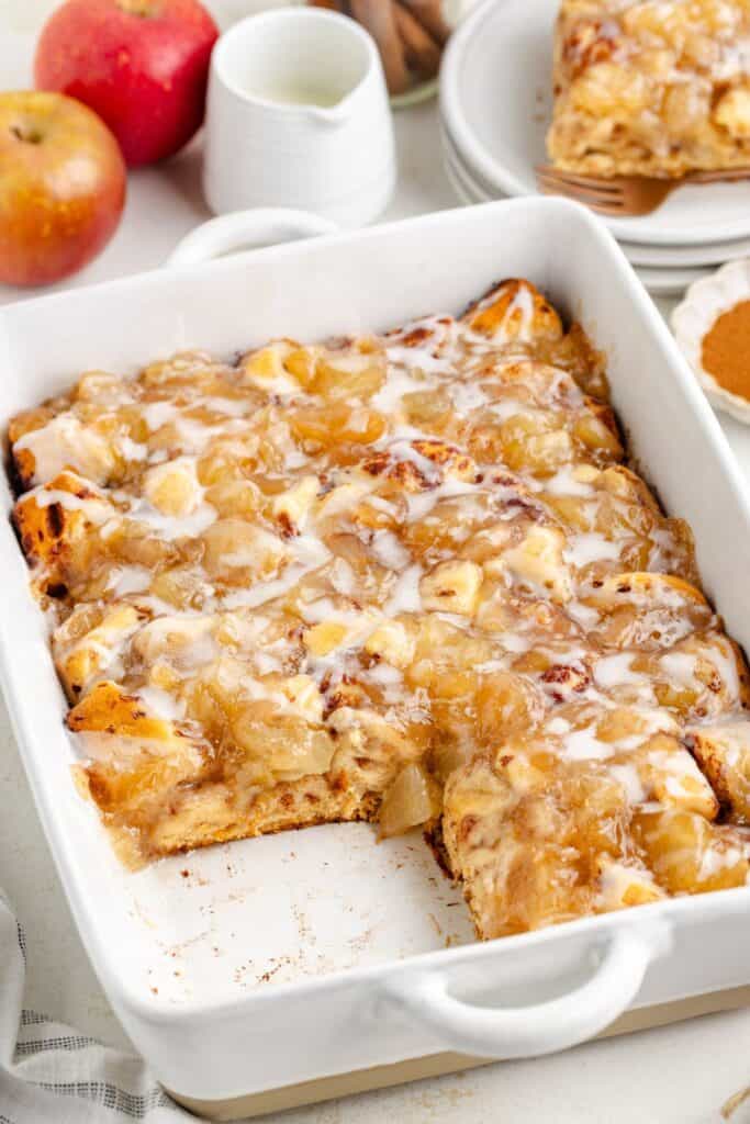 A baked apple cinnamon roll casserole with icing in a white dish; one piece missing, apples and plates behind.