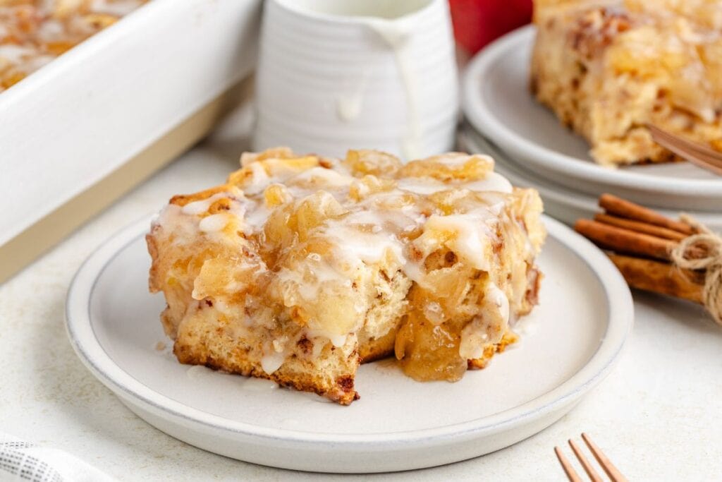 Slice of glazed cinnamon roll casserole with apple pie filling on a white plate, with cinnamon sticks and small pitcher behind.
