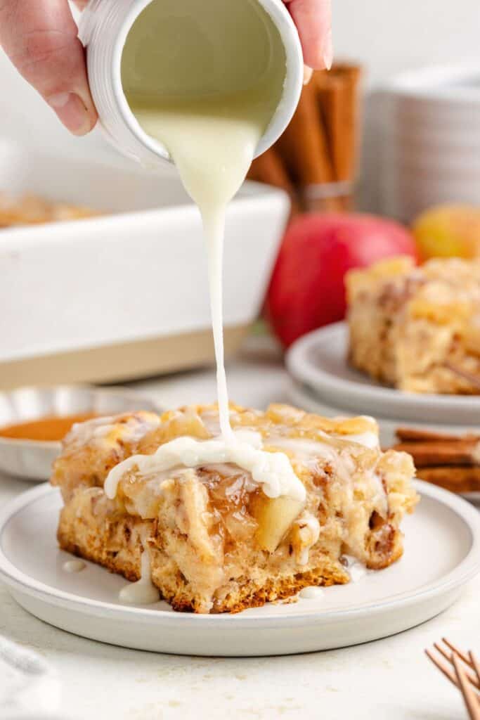 Hand pouring white icing onto apple cinnamon roll apple casserole on plate, with apples and cinnamon sticks behind.