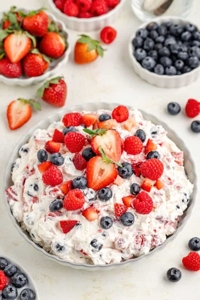 A bowl of mixed berry cheesecake salad topped with strawberries, blueberries, raspberries; fresh berries in background.