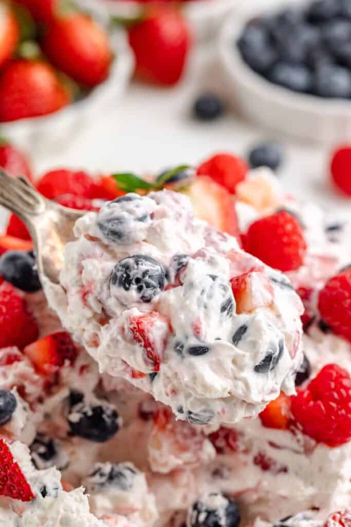 Close-up of a spoonful of cheesecake berry salad with strawberries, blueberries, and whipped cream; more berries behind.