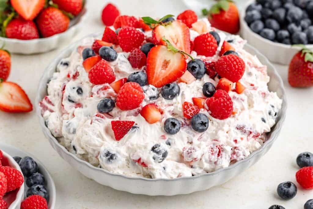 A bowl of creamy red, white, and blue cheesecake salad with fresh berries on top, surrounded by small dishes of berries.
