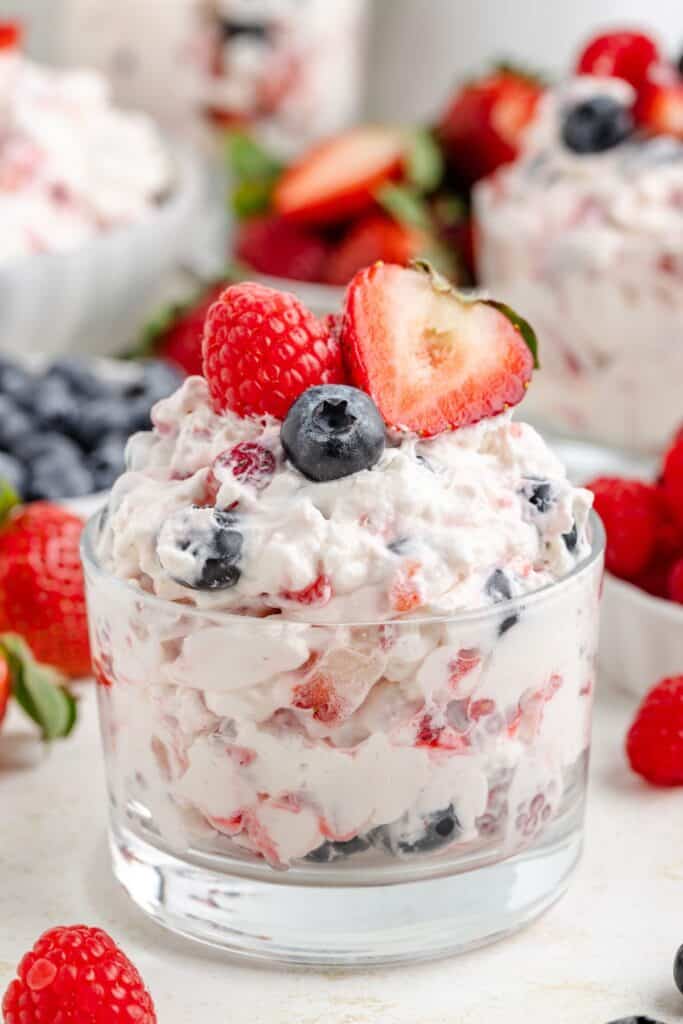 Glass cup of berry cheesecake dessert salad topped with fresh berries; extra berries and dessert bowls behind.