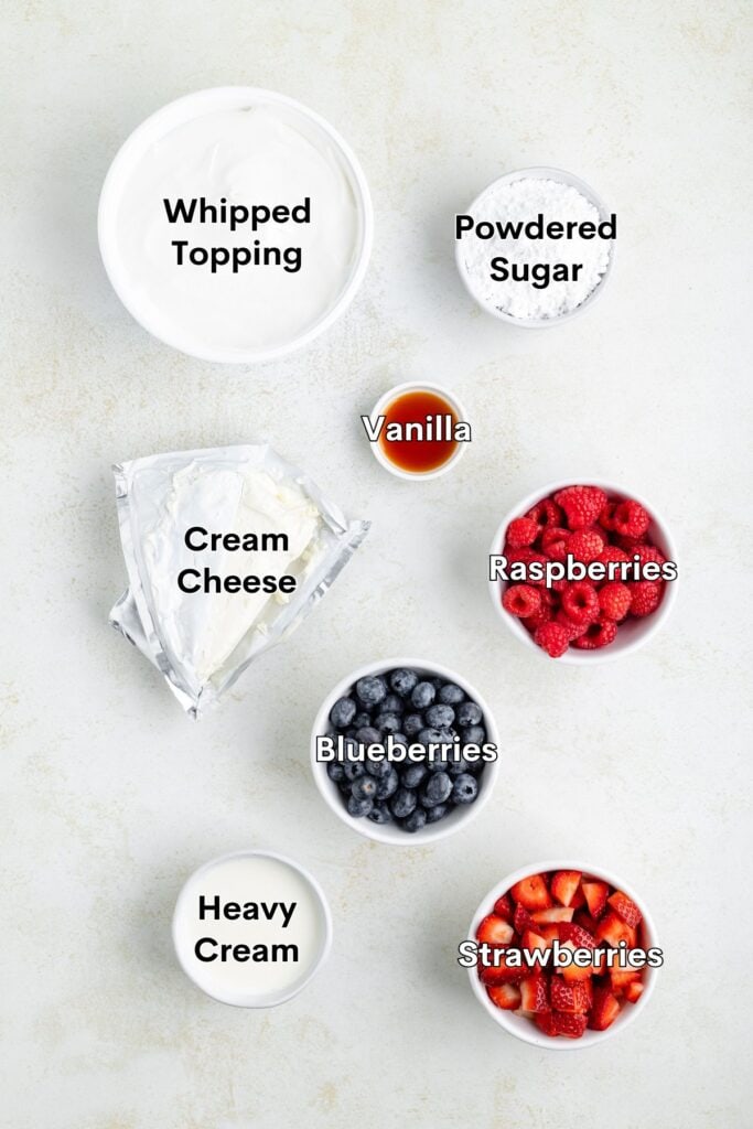 Assorted ingredients organized on a light background: berries, cream cheese, whipped topping, sugar, vanilla, and heavy cream.