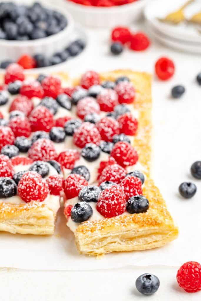 Rectangular puff pastry tart with cream, raspberries, blueberries, and powdered sugar on a white surface.