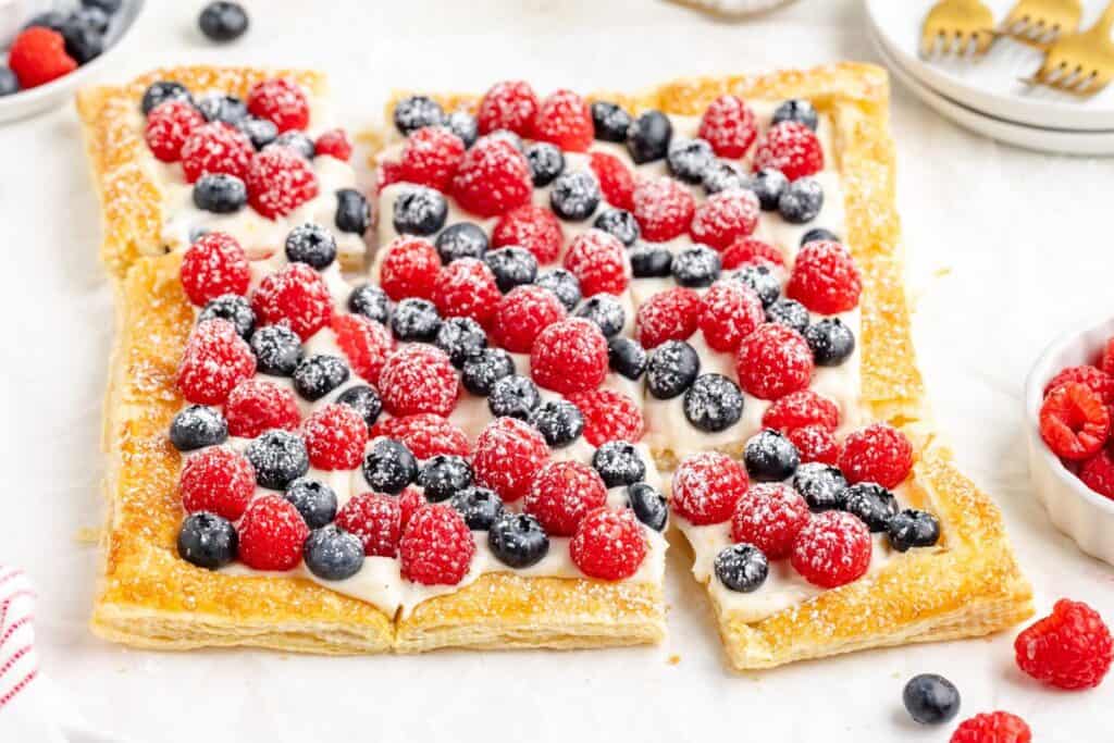 Rectangular puff pastry  tart topped with cream, raspberries, blueberries, powdered sugar; one slice slightly pulled out.