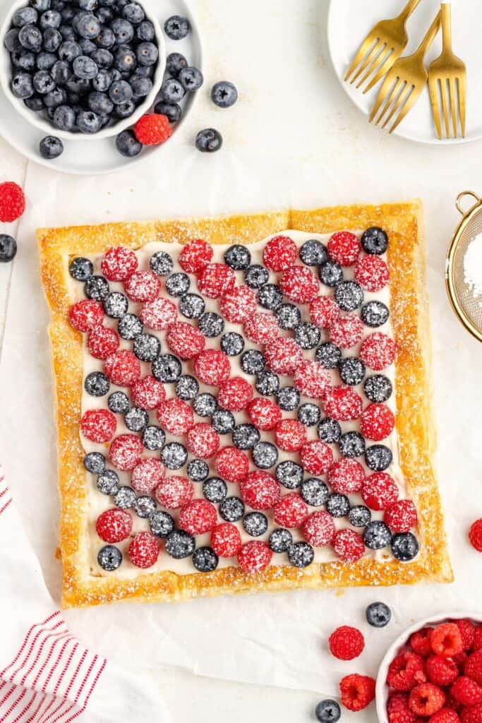 Square berry puff pastry  tart with raspberries, blueberries, powdered sugar; surrounded by bowls of berries and dessert forks.